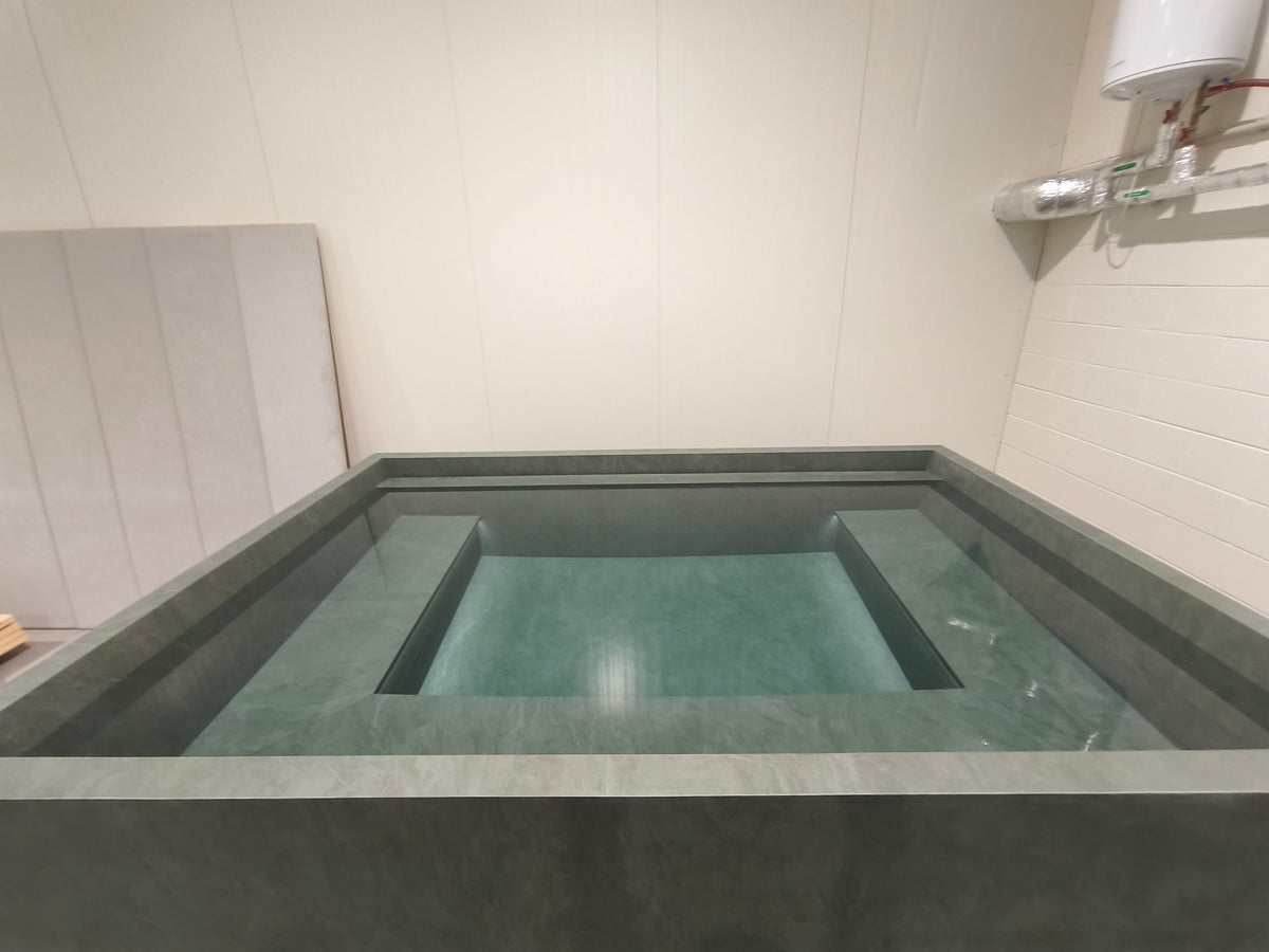 A modern rectangular outdoor hot spring or soaking tub with a dark gray stone exterior and a greenish-blue water-filled interior, set against a light beige tiled wall.