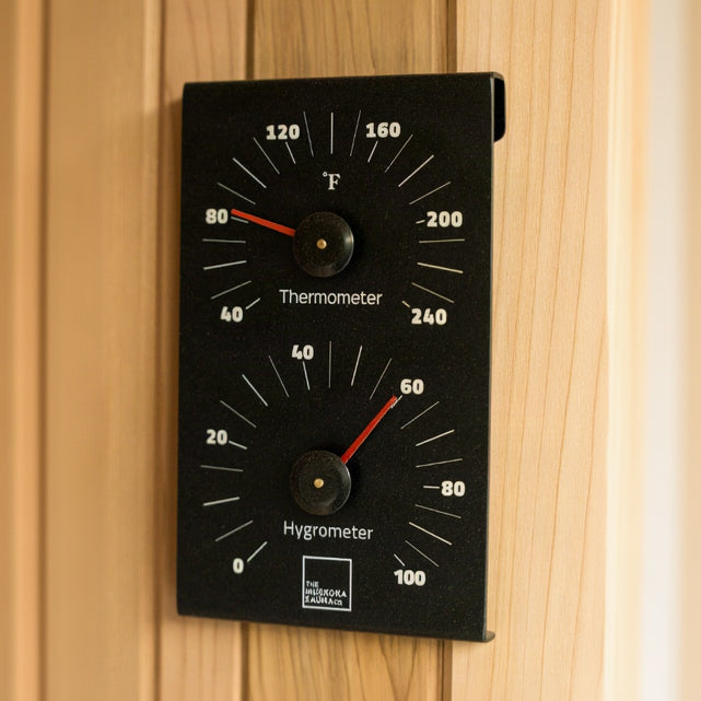 A black dual-panel wall-mounted thermometer and hygrometer with white markings and red needles, mounted on a light wood-paneled wall. The brand name 'The Muskoka Sauna Co.' is visible at the bottom of the hygrometer panel.